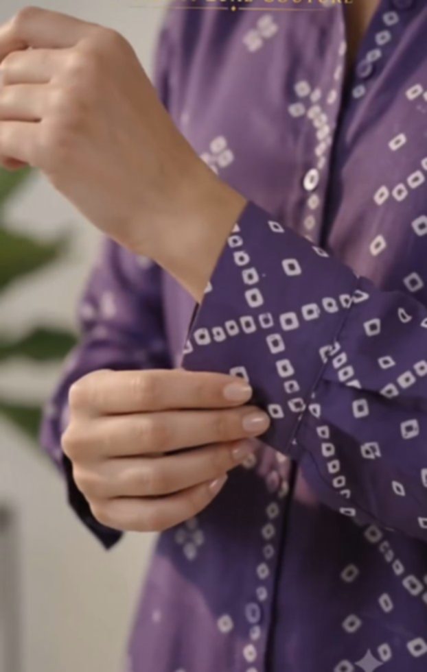Two hands fastening the button of a purple bandhani shirt, the small white circles of the tie-dye visible close up across the sleeve.