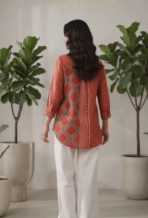 A woman seen from behind in a coral printed top with a geometric panel down the back, worn with white trousers, standing between two tall fig plants.