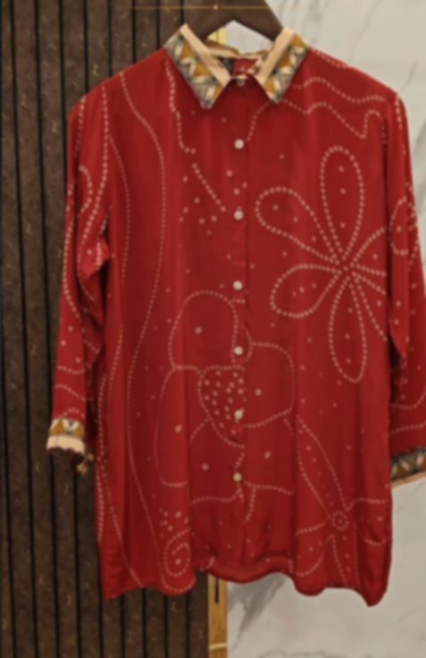 A deep red bandhani shirt on a hanger against dark wood, patterned with small white dots in a large flower, with an embroidered collar.
