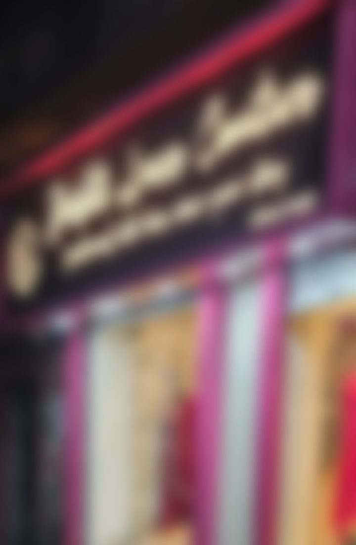 The shopfront at night. A black signboard carries Ashgrove Couture in lit gold script above the line fashioned with love, wear your story, with the name repeated in Bengali and the gold DLC monogram at the left. Below, magenta pillars frame a lit window with a red embroidered outfit on a mannequin, and a glass door.