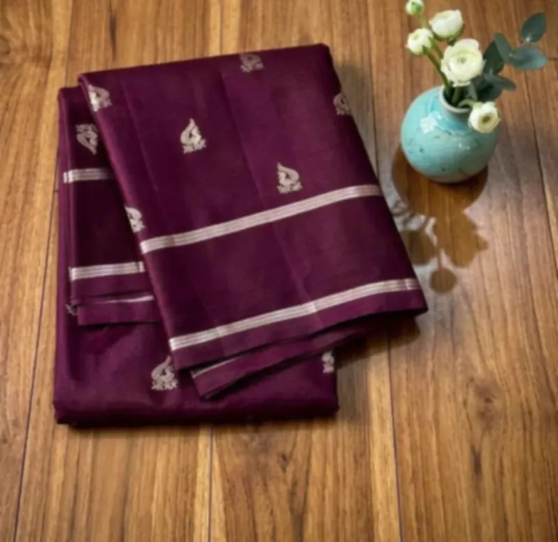 A deep aubergine silk saree folded on a walnut floor, with silver zari
                  motifs across it and two woven silver bands, beside a pale blue vase of
                  white ranunculus.