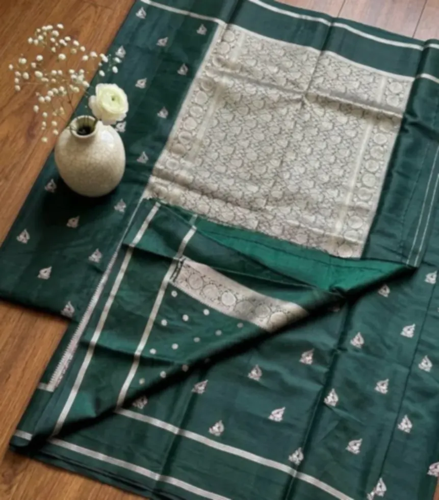 A deep green saree with a cream and silver brocade end, spread out on a wooden floor beside a stone vase of white flowers.
