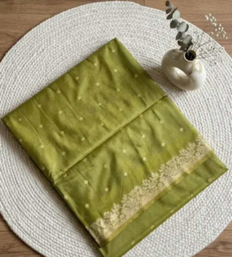 An olive green saree with small silver motifs and a gold border, folded on a round woven mat.