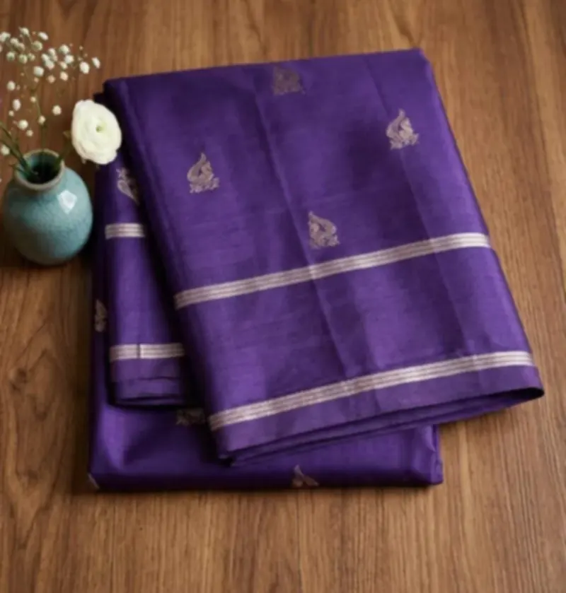 A violet saree with silver motifs and a silver border, folded on a wooden floor beside a vase of white flowers.