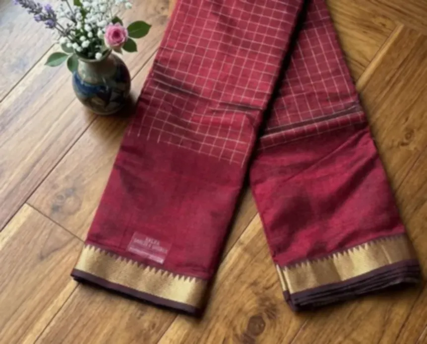 A wine red saree woven in a fine check, with a gold border, laid in a long fold on a wooden floor beside a painted vase of flowers.