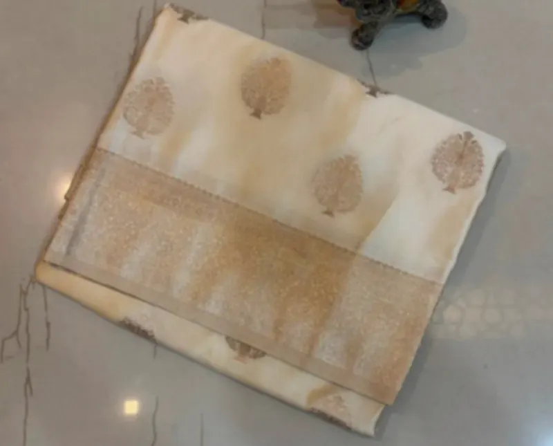 A cream silk saree with gold zari motifs and a wide gold border, folded on a pale floor.