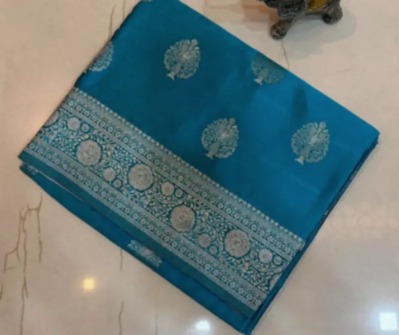 A peacock blue katan silk saree with silver zari motifs and a broad woven border, folded on a pale floor.