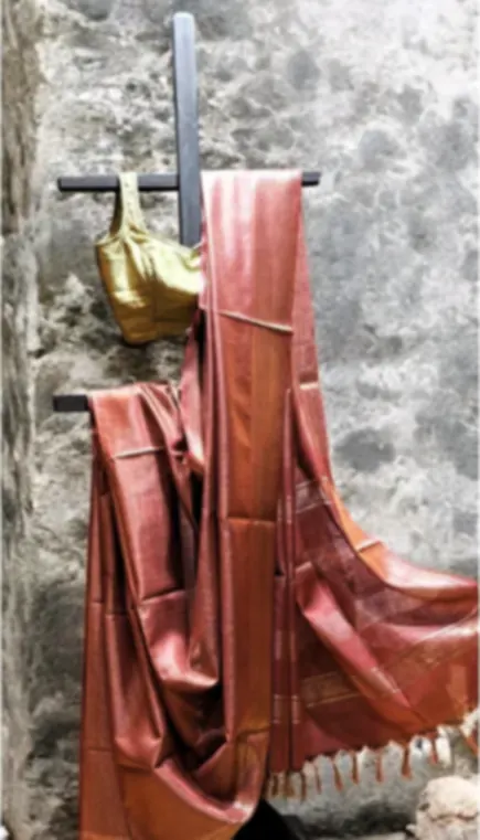 A rust Bhagalpuri blended silk saree draped over a rail against a stone wall, with a gold pot beside it.