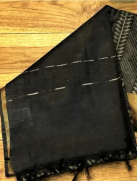 A black blended tussar saree with a gold zari border and fringe, laid on a wooden floor.