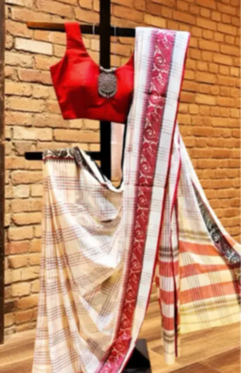 A cream Dhonekhali saree with red stripes, draped on a stand against a brick wall with a red blouse.