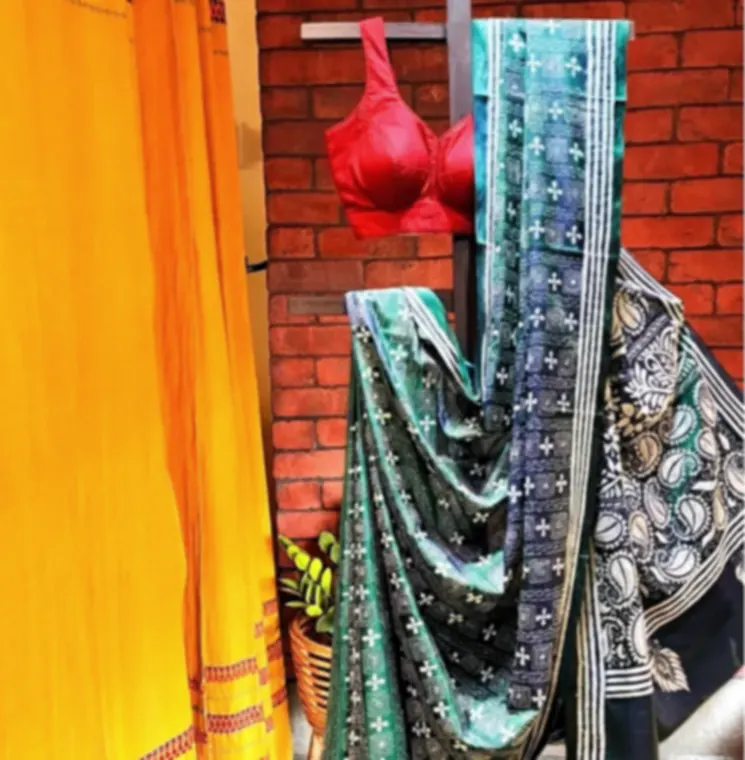 A teal blended silk saree with kantha embroidery, hanging in the shop beside a saffron drape and a red blouse.