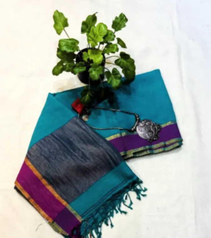 A teal handwoven khadi cotton saree with a purple and gold border, folded on white cloth with a silver pendant.