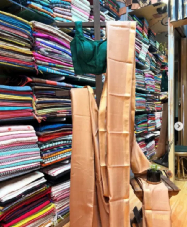 A peach Maheshwari saree with a gold zari border, hanging in the shop in front of stacked shelves of cloth.