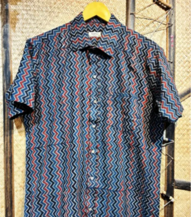 A blue and red Ajrakh printed cotton shirt with a zig-zag pattern, on a hanger against a woven backdrop.