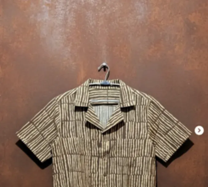 A striped pure cotton shirt with a soft collar, on a hanger against a rust-coloured wall.