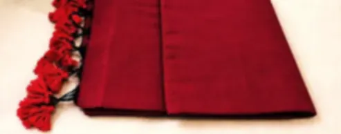 A deep red mulmul saree folded on cream cloth with a row of bold red tassels along its edge.