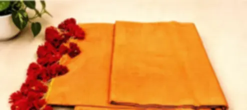 A warm orange mulmul saree folded on cream cloth with vivid red tassels along its edge.