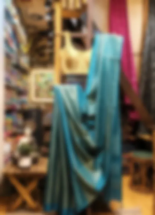 A blue silk blend saree draped on a display stand in the shop, with shelves of cloth behind it.