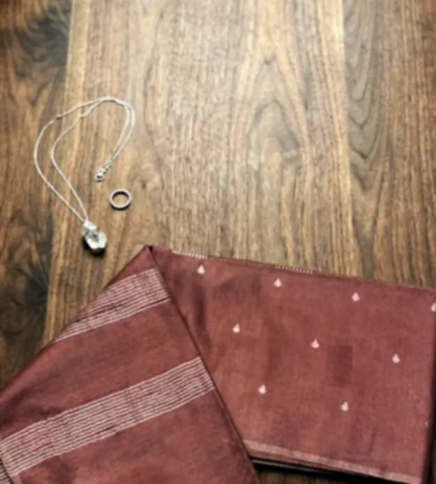 Two rosewood tissue sarees with fine silver stripes, folded on a dark wooden floor beside a silver chain.