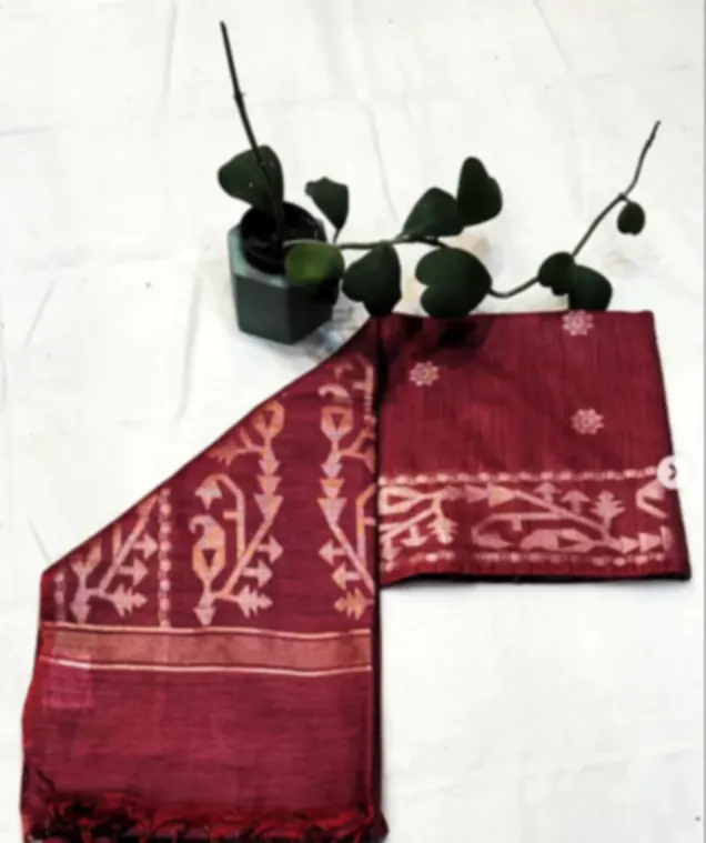 A maroon tussar saree woven with jamdani motifs, folded on white cloth with a small plant.