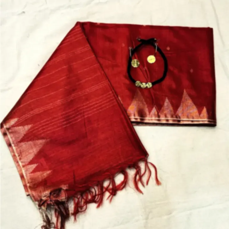 A red tussar saree with a woven temple border and fringe, folded on cream cloth beside a pendant.