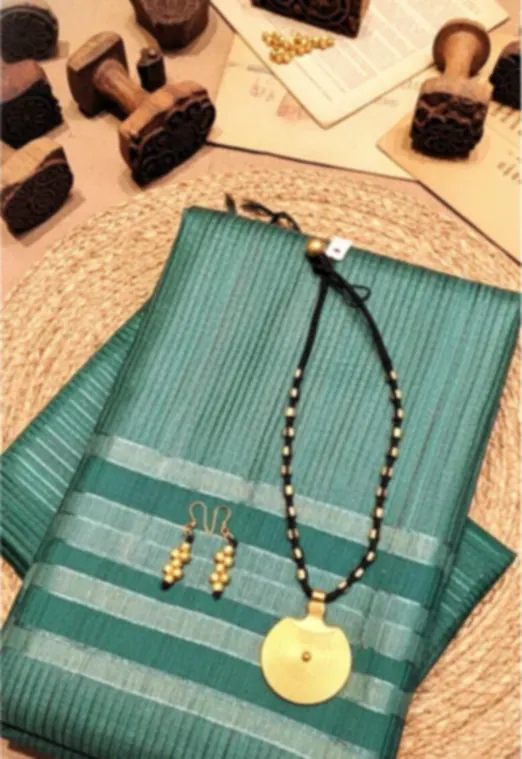 A teal tussar saree with woven stripes, folded on a round jute mat beside a brass pendant and printing blocks.