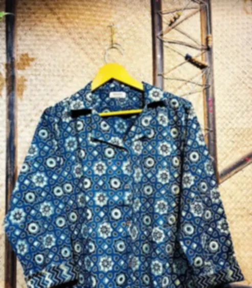 An indigo cotton shirt printed with Ajrakh medallions in white and blue, on a hanger against a textured wall.