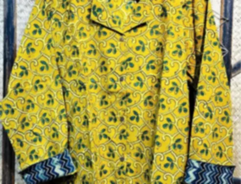 A mustard cotton shirt carrying Ajrakh lines and leaves, on a hanger against a woven backdrop.