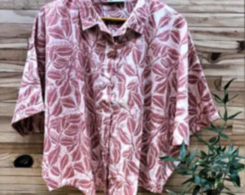 A pink cotton shirt with oversized foliage in muted terracotta, on a hanger beside a potted plant.