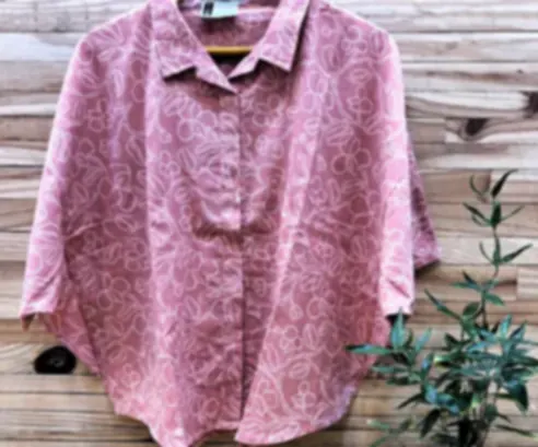 A dusty rose cotton shirt with loose hand-drawn florals, on a hanger beside a potted plant.